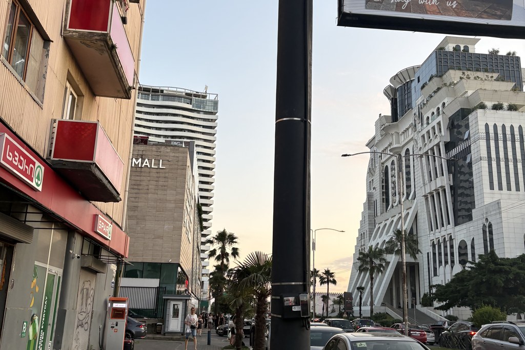 A Batumi street at dusk, mixing everyday shopfronts with modern towers and palm trees
